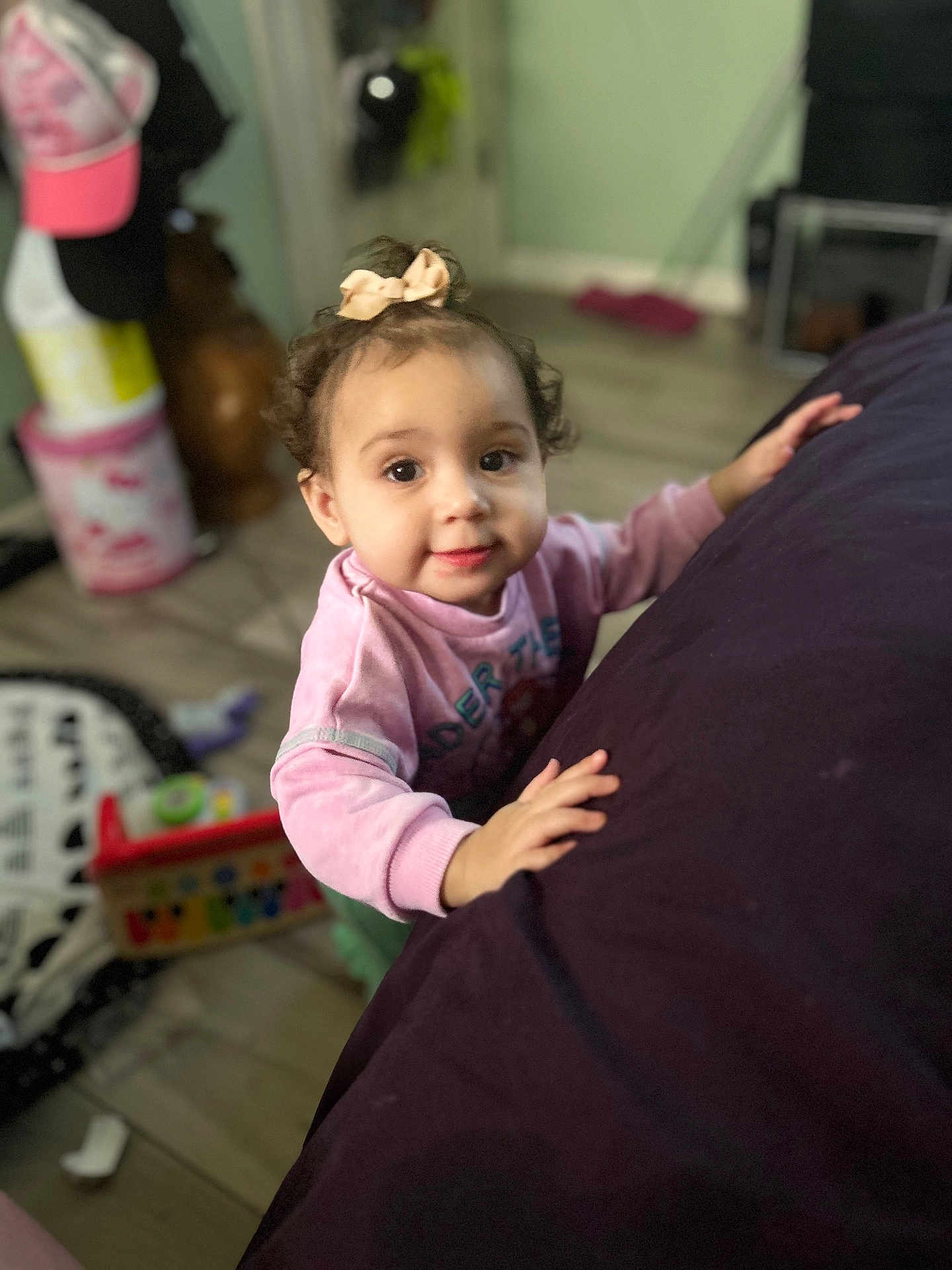 Stormie is registered to the contest to win money with this photo: toddler, child, indoor, curly_hair, pink_bow, pink_sweatshirt, toy, furniture, floor, wooden_floor, face, portrait, looking_up, hands, home, cozy, blurred_background, expression, young_child, casual