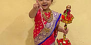 Meera is registered to the contest to win money with this photo: child, girl, traditional_clothing, saree, jewelry, smiling, pointing, barefoot, indoor, marble_floor, yellow_wall, decorated_staff, cultural, portrait, happy, cute, standing, person, festive, celebration
