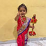 child, girl, traditional_clothing, saree, jewelry, smiling, pointing, barefoot, indoor, marble_floor, yellow_wall, decorated_staff, cultural, portrait, happy, cute, standing, person, festive, celebration
