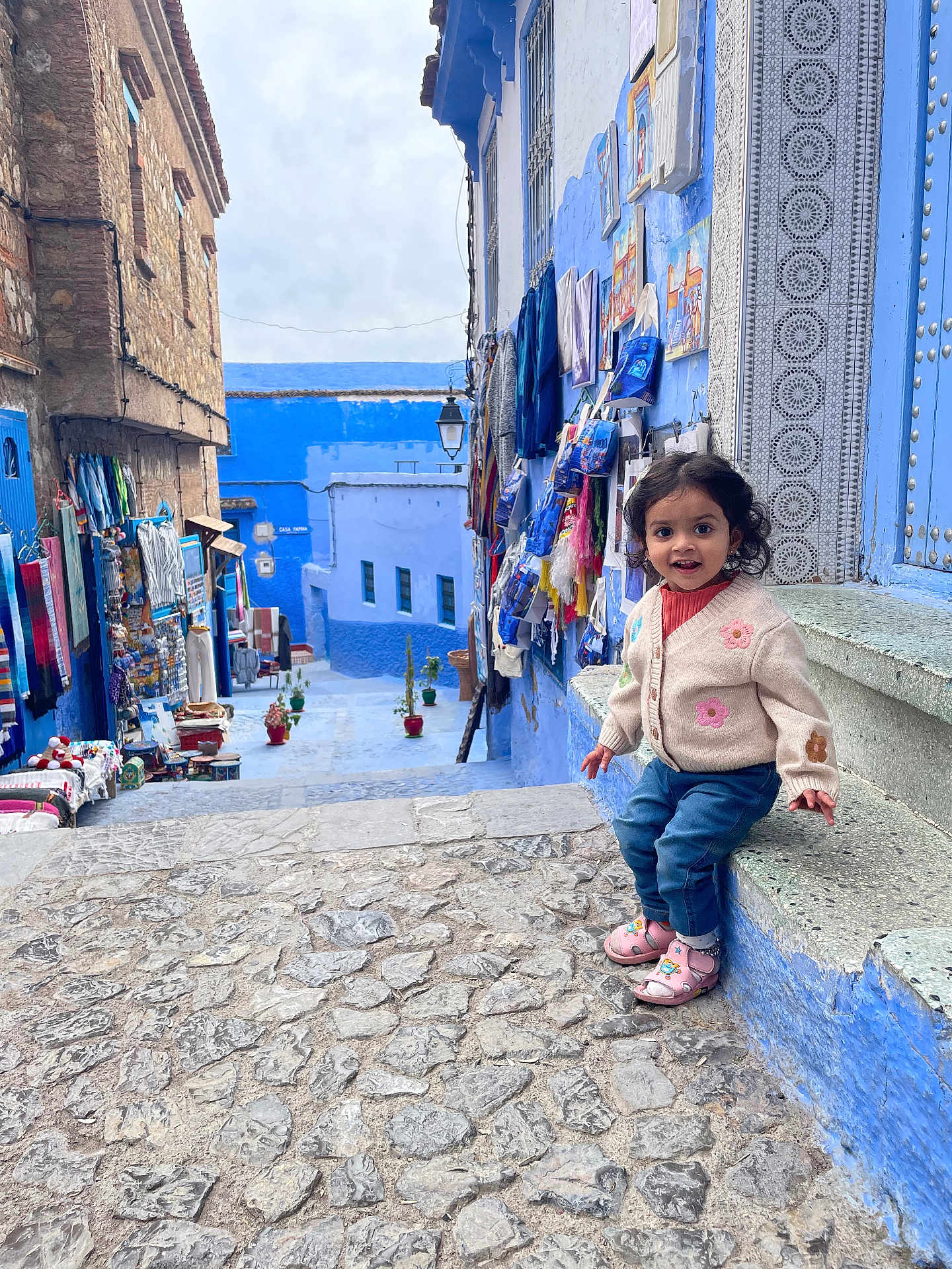 Meera joined the competition — help win amazing prizes! child, toddler, smiling, sweater, sandals, street, blue_walls, cobblestone, souvenirs, textiles, outdoor, daylight, person, window, building, lantern, curly_hair, stairs, colorful, travel