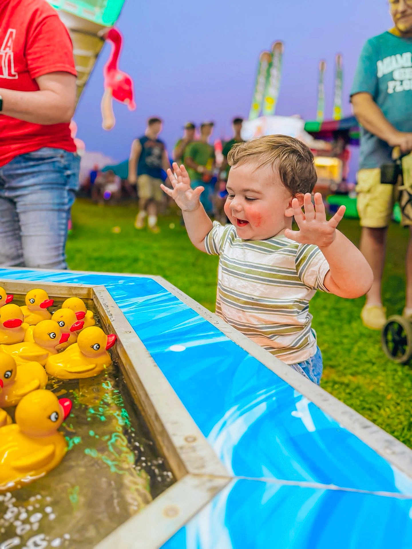 Brantlee is registered to the contest to win money with this photo: toddler, child, rubber_ducks, carnival_game, fairground, outdoor, grass, people, colorful, water, blue_surface, joyful, hands_up, smiling, casual_clothing, evening, fun, play, family_event, amusement_park