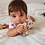 baby, bed, casual_clothing, child, close_up, curious, face, hand, indoor, large_eyes, lying_down, person, pink_toy, plush_toy, short_hair, skin, soft_light, teething_toy, toddler, white_bed
