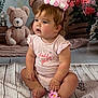 baby, child, girl, crown, headband, flower_bracelet, pink_clothing, teddy_bear, plush_toy, sitting, floor, wood_texture, curly_hair, cute, indoor, portrait, decor, soft_lighting, expression, adorable