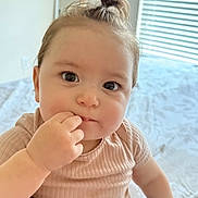 Yasmina is registered to the contest to win money with this photo: baby, child, infant, face, hand, fingers, hair, top_knot, clothing, pants, patterned_pants, bed, blanket, window, blinds, natural_light, indoors, cute, curious, sitting