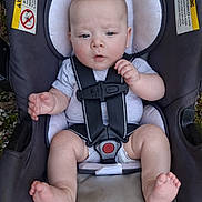 Waylon is registered to the contest to win money with this photo: baby, infant, child, car_seat, harness, portrait, sitting, indoors_outdoors, wide_eyed, facial_expression, hands, feet, bare_feet, gray_clothing, safety, vehicle_equipment, headrest, warning_label, grass, photography