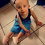 child, toddler, sitting, floor, tile, blue_shorts, pink_sneakers, person, legs, furniture, indoor, curious_expression, head_filter, casual_clothing, footwear, playing, home, light, shadow, looking_up