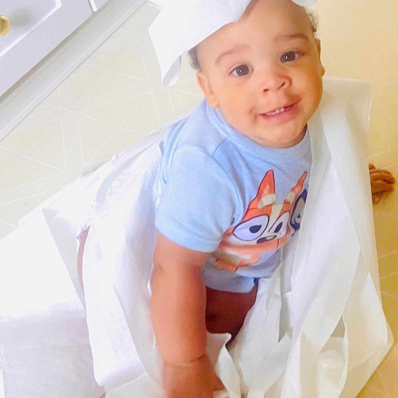 Christian joined the competition — help win amazing prizes! baby, blue_shirt, bright, casual_clothing, child, cute, floor, happy, indoor, infant, looking_up, one_child, paper_hat, person, playful, small_child, smiling, tile_floor, tissue_paper, white_cabinets