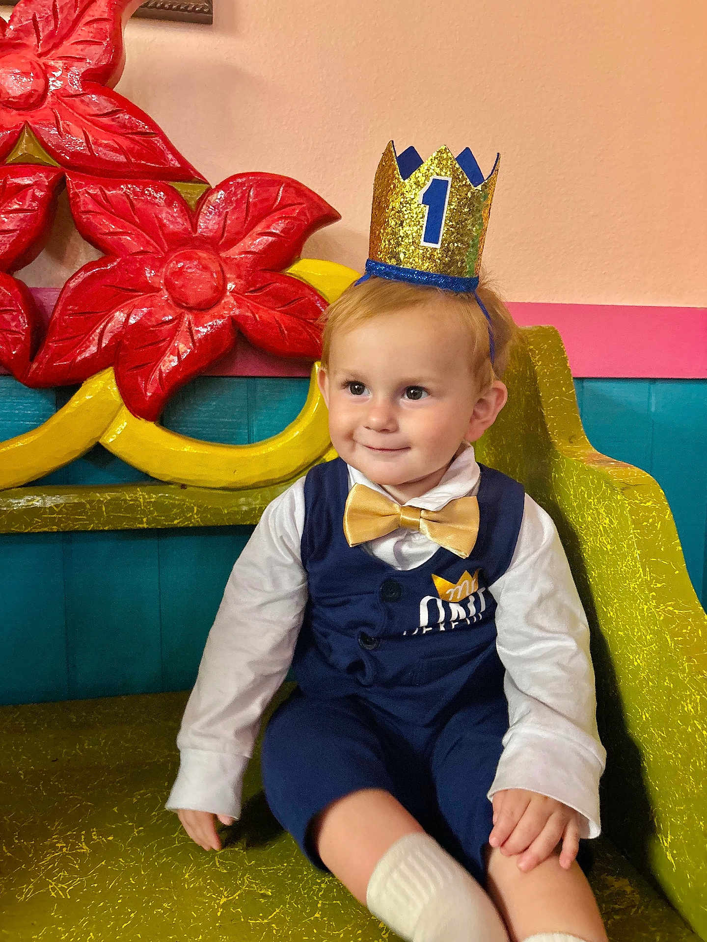 James is registered to the contest to win money with this photo: toddler, child, crown, birthday, party, golden_bow_tie, navy_vest, white_shirt, smile, bench, red_flowers, colorful_background, indoors, cute, celebration, happy, portrait, sitting, young_child, fashion