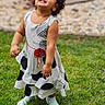 Kaeya joined the competition — help win amazing prizes! blurry_background, child, curly_hair, daylight, dress, family_photo, garden, girl, grass, happy, looking_up, outdoor, patterned_dress, playful, portrait, red_flower, shoes, smiling, standing, toddler