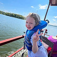 Kyah is registered to the contest to win money with this photo: child, life_jacket, peace_sign, boat, lake, water, outdoor, smiling, girl, summer, sunny, nature, trees, recreation, adventure, happy, person, blue_sky, water_splash, casual_clothing
