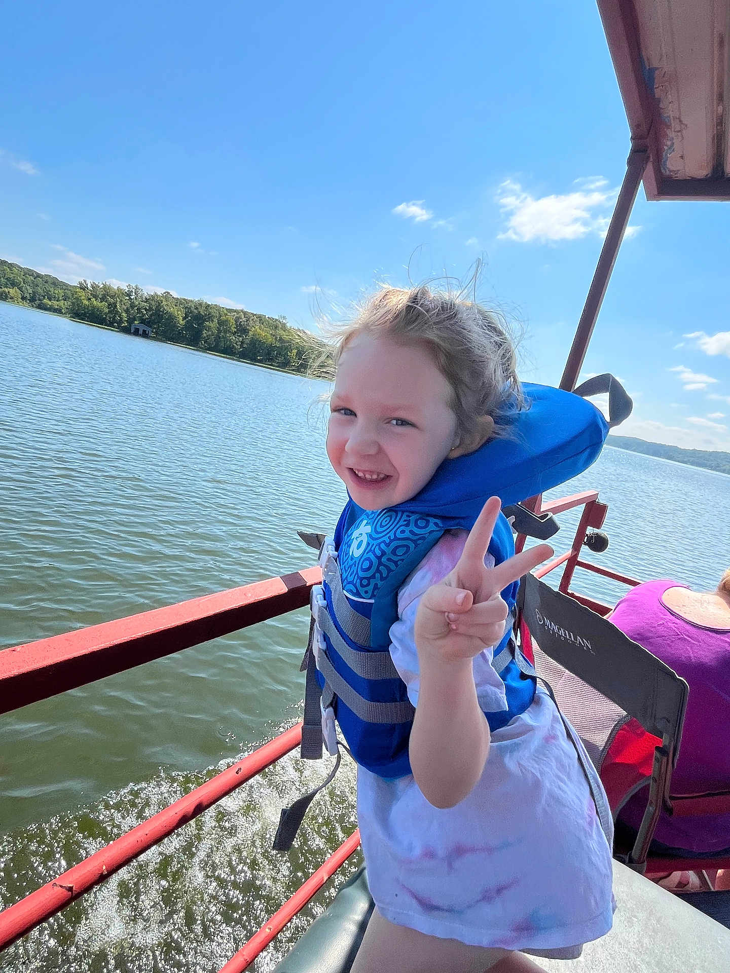 Kyah is registered to the contest to win money with this photo: child, life_jacket, peace_sign, boat, lake, water, outdoor, smiling, girl, summer, sunny, nature, trees, recreation, adventure, happy, person, blue_sky, water_splash, casual_clothing