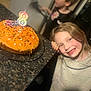 birthday_cake, candle, child, smile, happy, party, celebration, cake, sprinkles, kitchen_counter, girl, blond_hair, casual_clothing, indoor, family, fun, festive, food, young, portrait