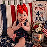 baby, child, red_bow, chair, american_flag, blanket, flowers, lantern, patriotic, celebration, holiday, decorations, sign, smile, indoors, portrait, cute, festive, fourth_of_july, happy