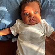 Kehlani joined the competition — help win amazing prizes! baby, infant, onesie, white_clothing, bed, blue_bedspread, brown_bedspread, curly_hair, face, wide_eyes, sunlight, expression, laying_down, child, person, cute, indoors, portrait, skin, soft_light