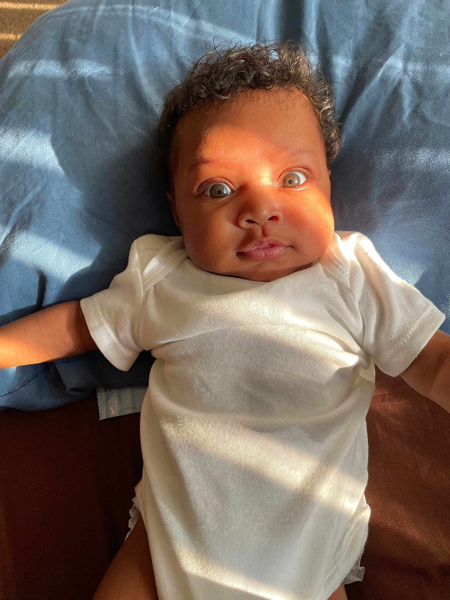 Kehlani joined the competition — help win amazing prizes! baby, infant, onesie, white_clothing, bed, blue_bedspread, brown_bedspread, curly_hair, face, wide_eyes, sunlight, expression, laying_down, child, person, cute, indoors, portrait, skin, soft_light