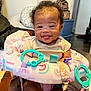 Penelope is registered to the contest to win money with this photo: baby, smiling, tongue_out, infant, baby_chair, toy, indoor, happy, cute, child, person, home, decor, seated, clothing, curly_hair, playful, colorful, comfort, portrait