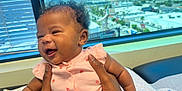 Penelope is registered to the contest to win money with this photo: baby, infant, smiling, holding_hands, pink_clothing, curly_hair, indoor, window, cityscape, daylight, person, skin, arm, happy, cute, child, expression, face, seated, table