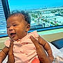 baby, infant, smiling, holding_hands, pink_clothing, curly_hair, indoor, window, cityscape, daylight, person, skin, arm, happy, cute, child, expression, face, seated, table