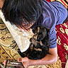 person, puppy, dog, phone, pillow, rug, carpet, lying_down, indoor, hand, black_hair, blue_shirt, cute, pet, relaxing, snuggling, resting, cozy, home, casual