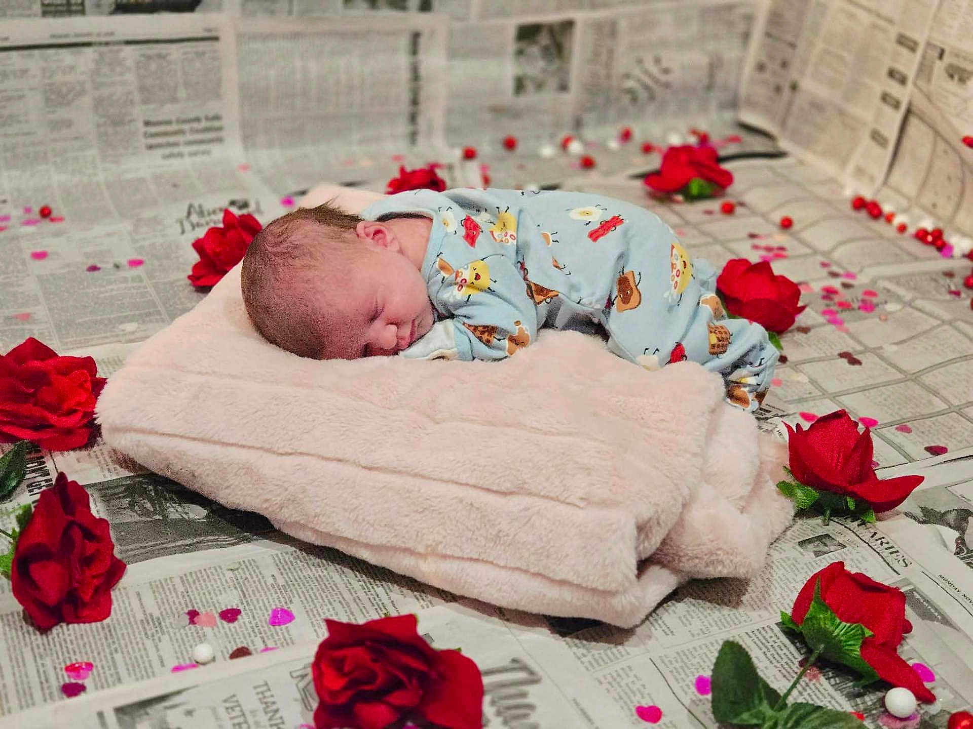 Zoey is registered to the contest to win money with this photo: baby, sleeping, blanket, onesie, red_roses, confetti, newspaper, soft_texture, cute, infant, peaceful, lying_down, decorative, indoors, portrait, child, resting, floral, celebration, cozy
