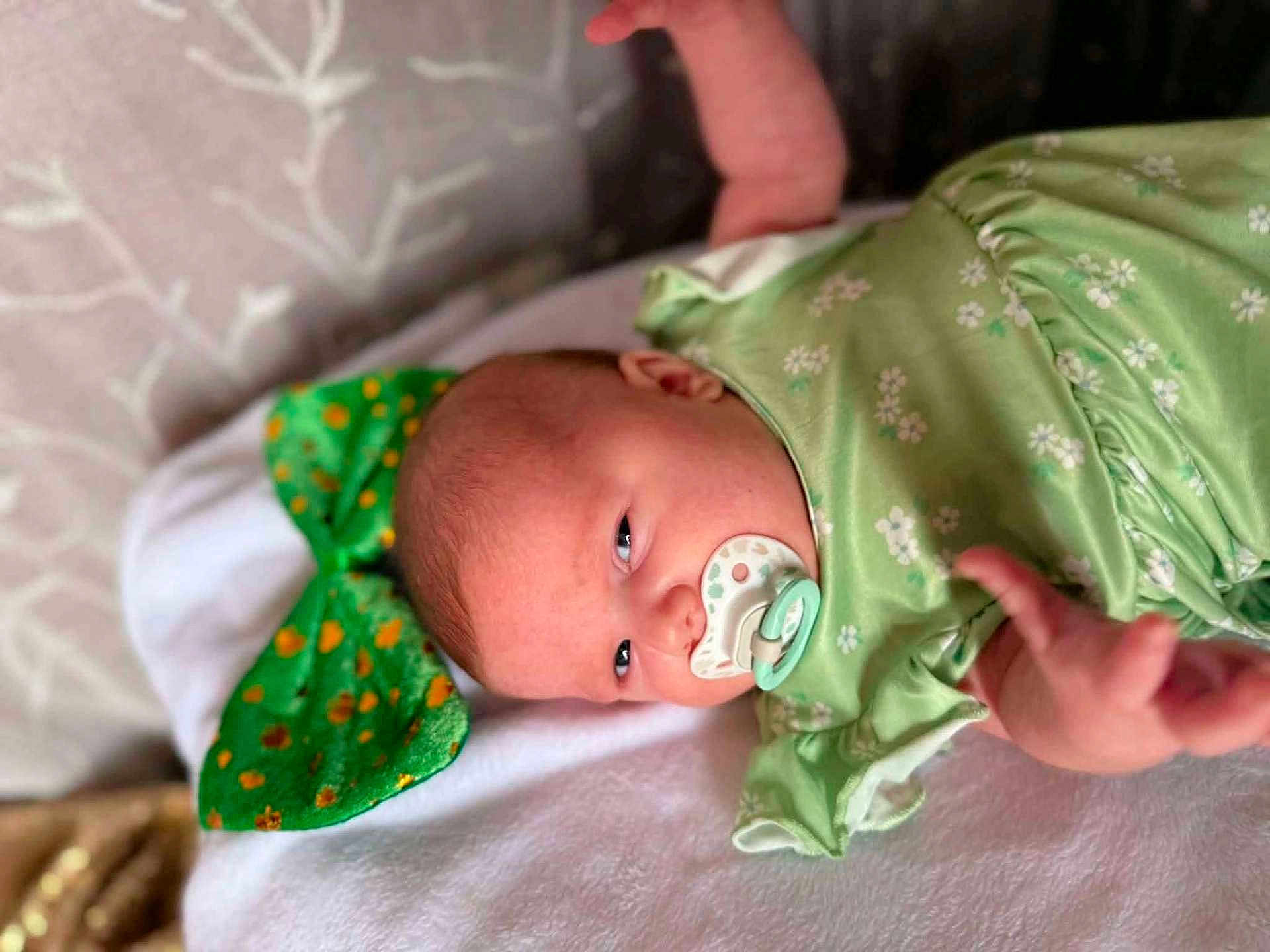 Zoey is registered to the contest to win money with this photo: baby, infant, newborn, pacifier, green_outfit, floral_pattern, lying_down, blanket, bedding, head, face, eyes_open, hand, pillow, bow, soft_texture, portrait, cute, nursery, skin