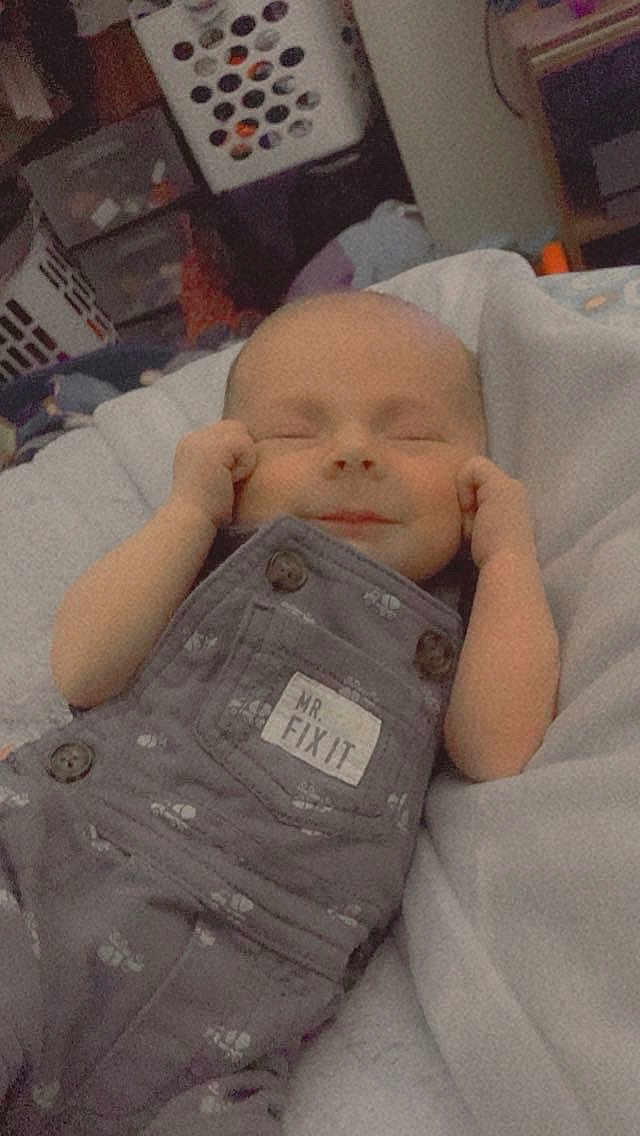 Carson is registered to the contest to win money with this photo: baby, infant, newborn, smiling, sleeping, overalls, onesie, pocket_label, text_on_clothing, hands, face, bed, blanket, nursery, laundry_basket, furniture, indoors, cute, peaceful, portrait