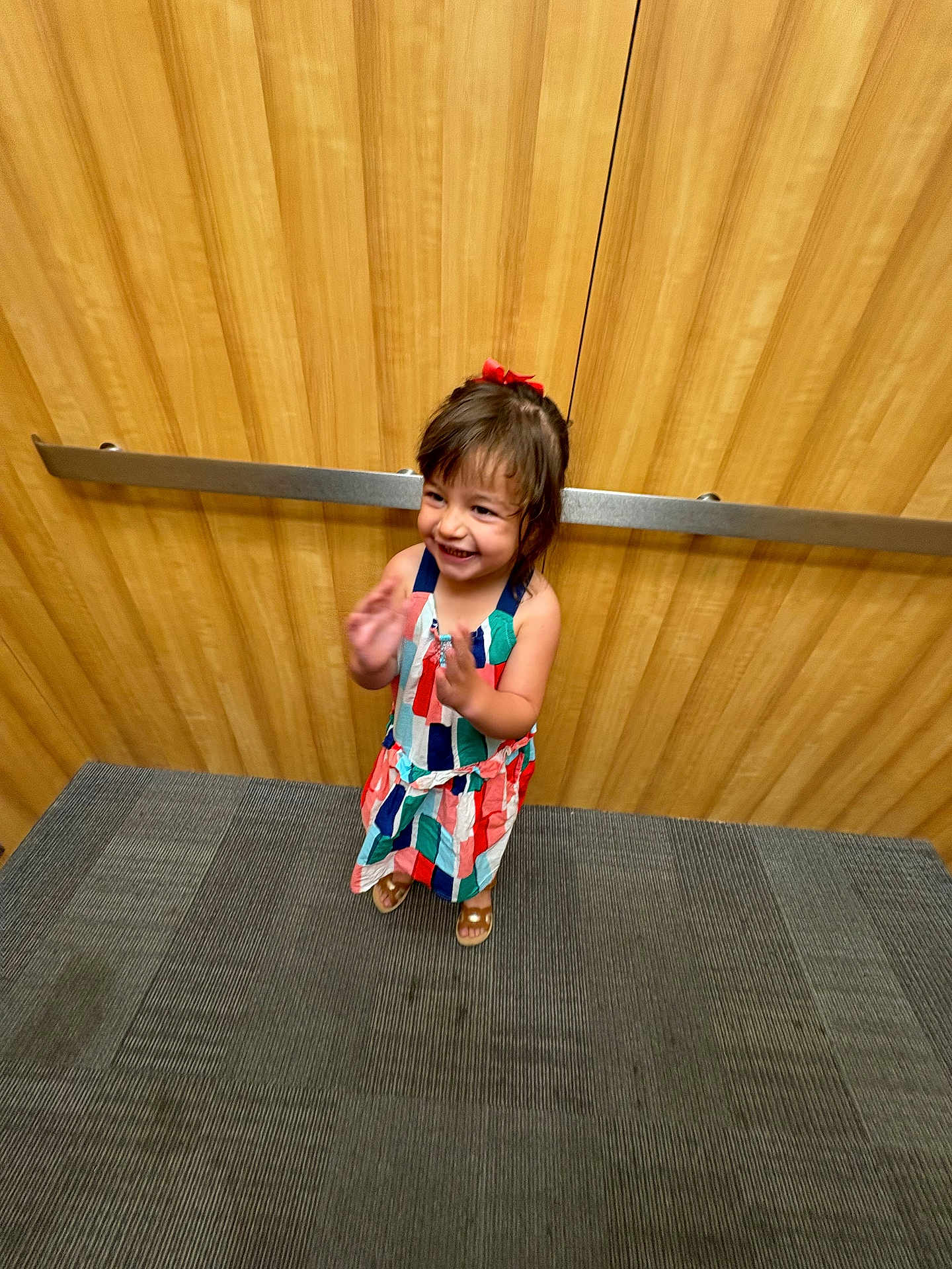 Cora is registered to the contest to win money with this photo: child, girl, smiling, clapping, elevator, wood_paneling, carpet, red_bow, dress, sandals, happy, indoors, young_child, person, cute, standing, joyful, playful, colorful_clothing, hair