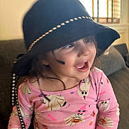Cora is registered to the contest to win money with this photo: toddler, child, hat, black_hat, pink_shirt, ghost_pattern, smiling, happy, indoor, couch, window, blinds, long_sleeve, face, hair, playful, cute, expression, clothing, person