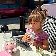 Cora is registered to the contest to win money with this photo: car, chair, child, daytime, drinking, focus, girl, glass, hair, messy_ponytail, outdoor, pacifier, pink_clothing, plush, pouch, street, sunlight, table, tablet, toy