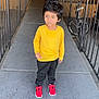child, boy, yellow_shirt, black_pants, red_sneakers, concrete, walkway, metal_railings, door, bicycle, outdoor, standing, hair, blue_eyes, expression, casual_clothing, urban, daylight, side_view, person