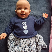 Elvira participe au concours pour gagner de l'argent avec cette photo : baby, child, smiling, blue_blanket, navy_top, heart_design, patterned_skirt, lying_down, happy, cute, infant, person, indoors, soft_texture, toddler, clothing, portrait, face, head, small_hands