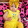 baby, child, yellow_clothing, headband, bow, sitting, cushion, pink_pattern, black_pattern, indoor, cute, portrait, infant, person, fashion, colorful, expression, eyes, hand, footwear