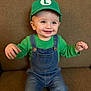 baby, cap, casual, child, clothing, couch, cute, denim_overalls, face, green_hat, green_shirt, happy, headwear, indoors, person, portrait, sitting, smiling, toddler, young_child