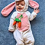 baby, bunny_costume, pink_ears, carrot_toy, blue_blanket, smiling, waving_hand, cute, infant, soft_texture, lying_down, child, hooded_outfit, plush, happy_face, costume, indoors, person, portrait, cozy