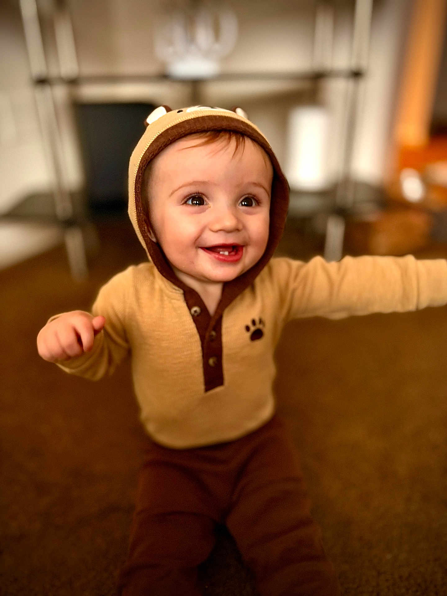 Mazen is registered to the contest to win money with this photo: baby, child, toddler, smiling, happy, hood, ears, brown_clothing, beige_clothing, carpet, indoor, floor, person, cute, portrait, arms_outstretched, blurred_background, home, sitting, infant
