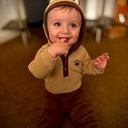 Mazen is registered to the contest to win money with this photo: baby, child, smiling, hoodie, brown_clothing, sitting, carpet, indoor, cute, happy, face, finger_in_mouth, cozy, young_child, portrait, infant, warm_colors, playful, person, soft_focus