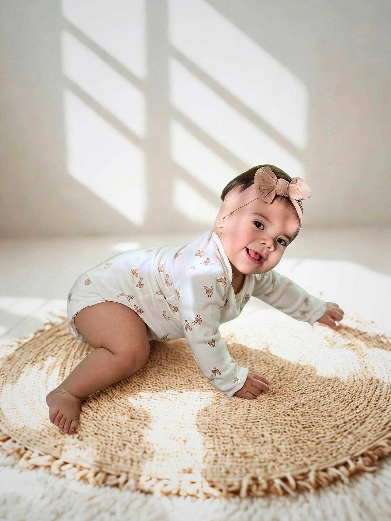 Aurora is registered to the contest to win money with this photo: baby, infant, crawling, onesie, headband, bow, smiling, happy, cute, indoor, rug, woven_rug, natural_light, shadow, playful, child, person, floor, home, soft_light