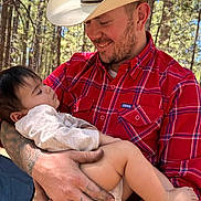 Aurora joined the competition — help win amazing prizes! man, baby, child, forest, trees, hat, cowboy_hat, plaid_shirt, red_shirt, tattoos, watch, outdoor, nature, holding, smiling, peaceful, sleeping, baby_clothes, arm, daylight