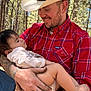 man, baby, child, forest, trees, hat, cowboy_hat, plaid_shirt, red_shirt, tattoos, watch, outdoor, nature, holding, smiling, peaceful, sleeping, baby_clothes, arm, daylight
