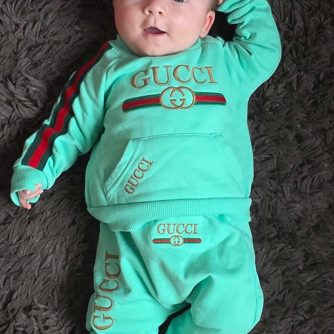 Zélie a rejoint le concours — aidez-le/la à gagner de superbes lots ! baby, blue_eyes, branded_clothing, carpet, child, cute, face, fur_rug, gucci_logo, hand, human, indoors, infant, lying_down, portrait, smile, socks, soft_texture, sweatpants, sweatshirt