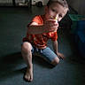 barefoot, blurry, boy, candid, carpet, casual, child, denim_shorts, face, floor, hand, indoor, kneeling, low_light, orange_shirt, playful, portrait, room, toy, young