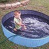 Paul is registered to the contest to win money with this photo: child, pool, water, grass, backyard, tree, hose, stone_border, outdoor, summer, greenery, casual_clothing, boy, sitting, reflection, small_pool, daytime, nature, play, relaxation