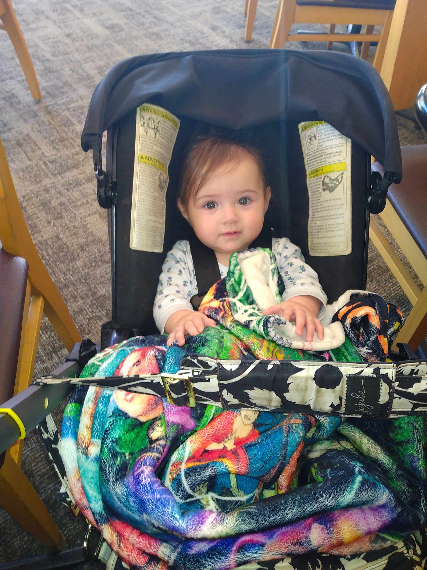 Bella is registered to the contest to win money with this photo: baby, infant, stroller, car_seat, blanket, colorful_blanket, seatbelt, hands, eyes, hair, face, indoor, chair, carpet, floor, buckle, clothing, portrait, calm_expression, hood/canopy