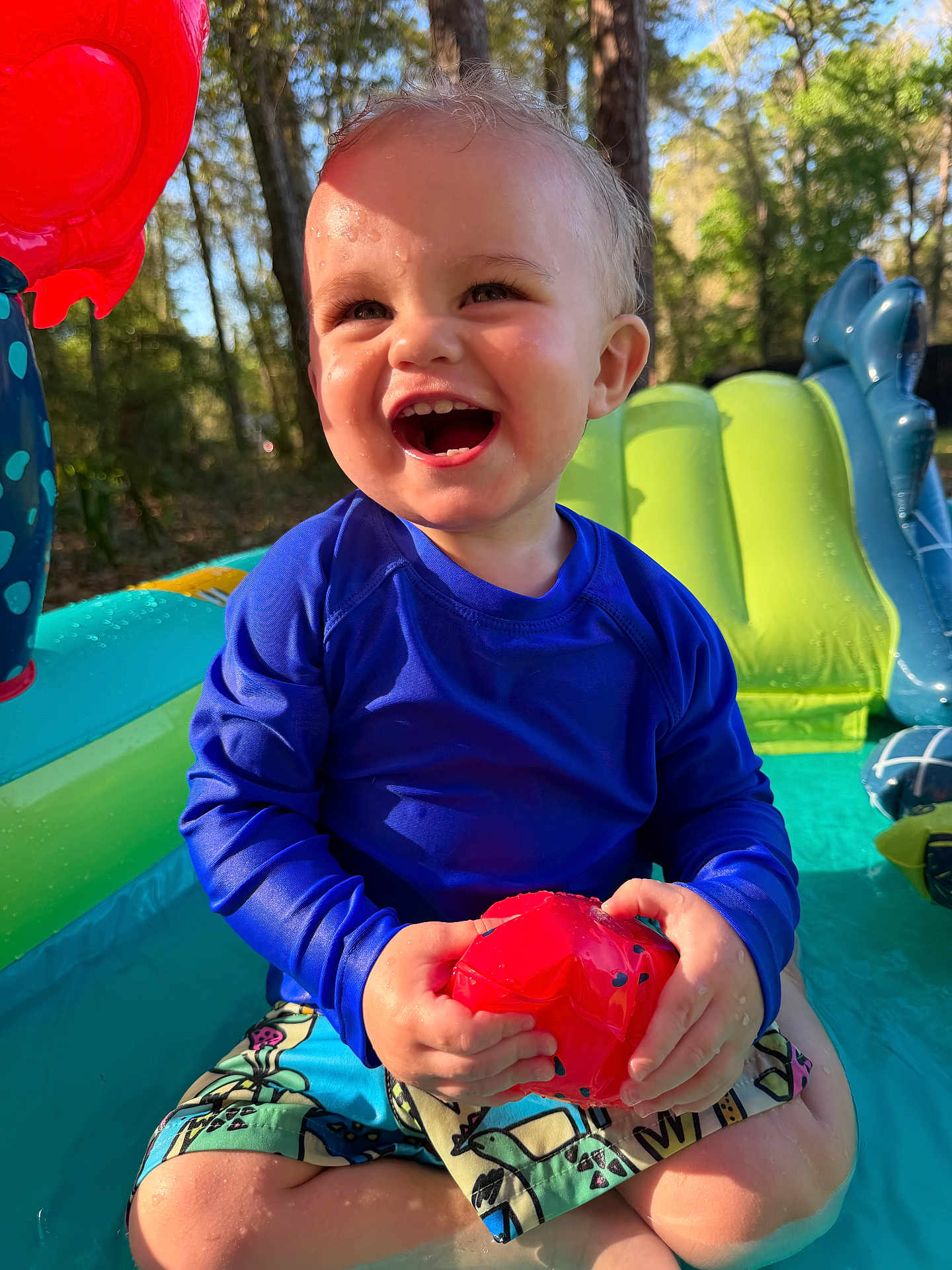 Hudson is registered to the contest to win money with this photo: child, toddler, smile, laughing, inflatable_pool, swim_shirt, swim_trunks, red_toy, water, wet_hair, outdoors, trees, sunlight, playtime, backyard, close_up, joyful, sitting, summer, skin