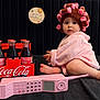 baby, pink_robe, hair_curlers, vintage_phone, coca_cola, glass_bottles, black_background, sign, 10_month, child, curious, sitting, toy, retro, portrait, indoors, cute, wide_eyes, floor, hat