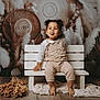 artistic_background, barefoot, bench, child, cozy, curly_hair, cute, decor, dried_flowers, indoor, long_sleeve_shirt, overalls, playful, portrait, smile, texture, toddler, tongue_out, white_bench, wooden_floor