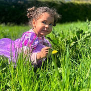 Lena a rejoint le concours — aidez-le/la à gagner de superbes lots ! child, toddler, purple_dress, grass, flower, dandelion, outdoor, nature, greenery, sunlight, curly_hair, garden, playful, crouching, daylight, smiling, person, portrait, young_child, happy