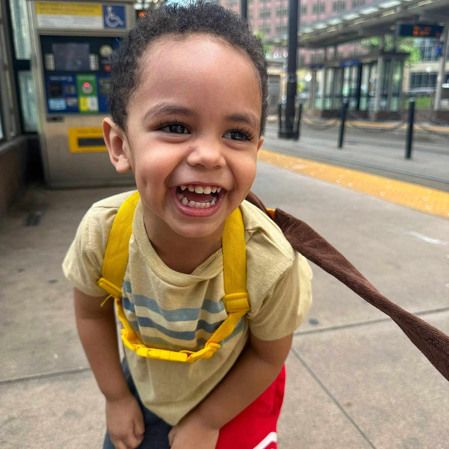 Willdrian joined the competition — help win amazing prizes! candid, casual_clothing, child, concrete, curly_hair, daylight, dimples, joy, laughing, playful, portrait, public_transport, red_shorts, shoes, sidewalk, smile, toddler, transit_station, urban, yellow_backpack