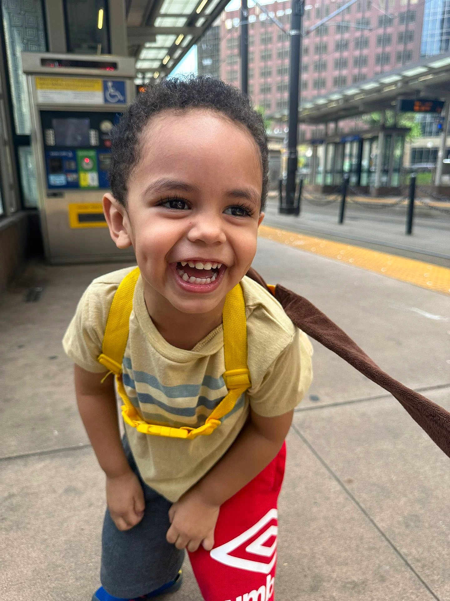 Willdrian joined the competition — help win amazing prizes! candid, casual_clothing, child, concrete, curly_hair, daylight, dimples, joy, laughing, playful, portrait, public_transport, red_shorts, shoes, sidewalk, smile, toddler, transit_station, urban, yellow_backpack