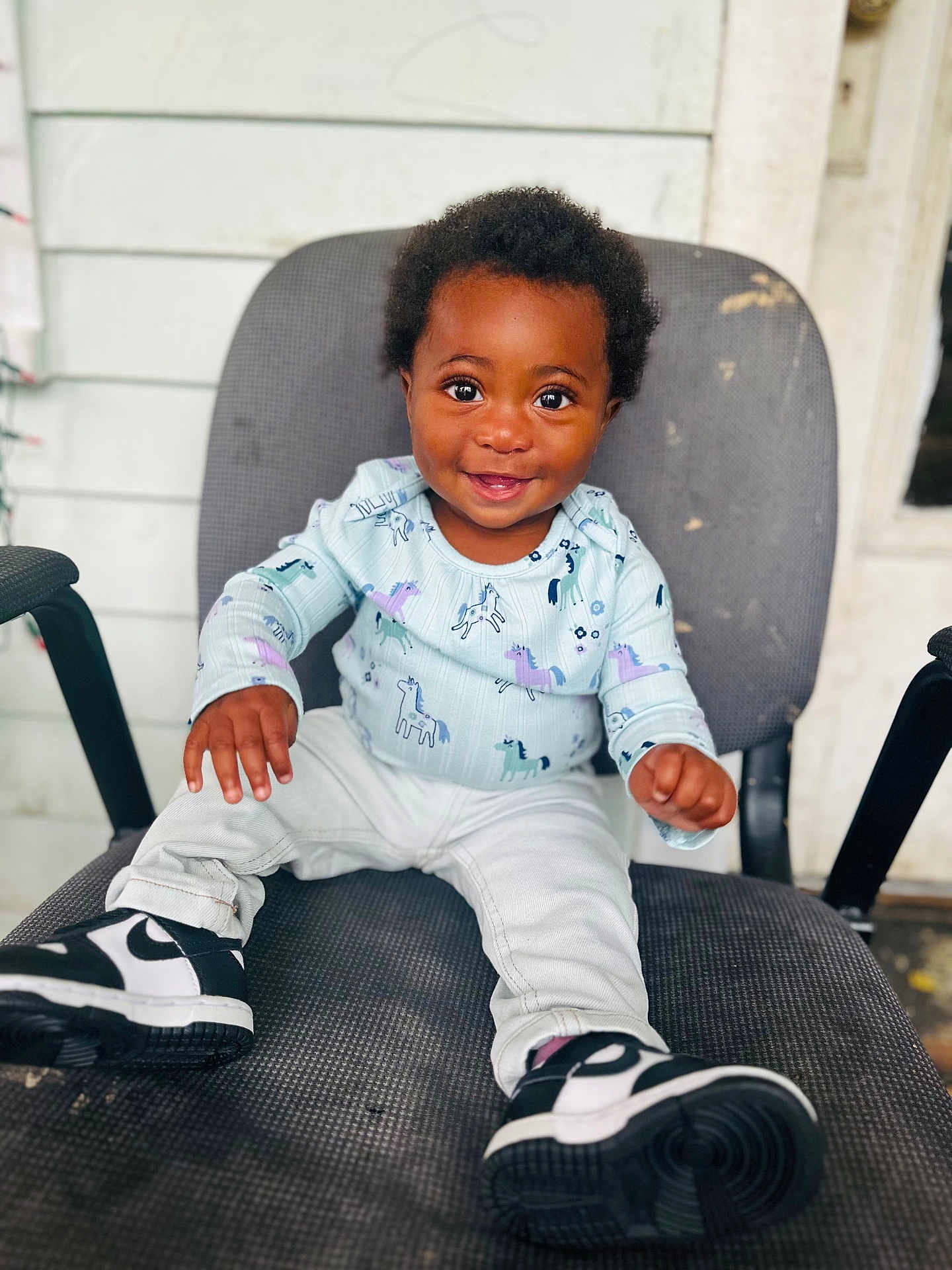 Jahmira is registered to the contest to win money with this photo: black_hair, chair, child, clothing, cute, dark_skin, eyes, feet, hands, happy, indoors, person, portrait, shirt, sitting, smiling, sneakers, toddler, unicorn_pattern, white_pants